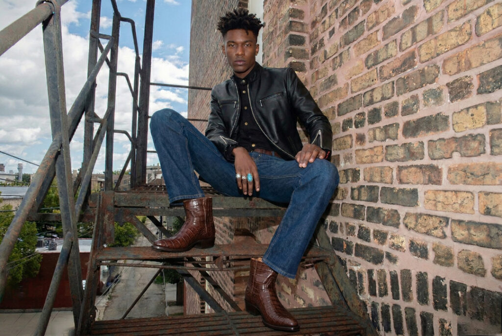 A young Black man wearing a White Widow Leather black moto jacket and brown alligator-pattern leather boots, sitting on a rusty fire escape against a brick wall.